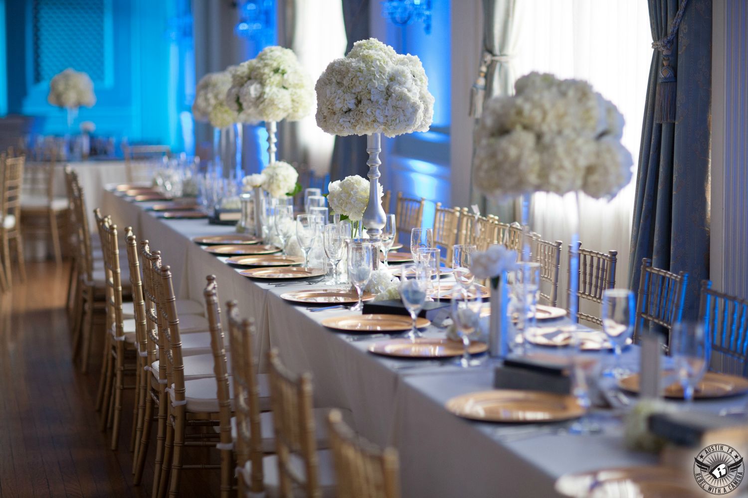 Stunningly decorated wedding reception with gold chiavari chairs, white hydrangeas in silver vases, and gold chargers with blue uplighting in the ballroom at the Texas Federation of Women's Clubs Mansion Headquarters decor by Clearly Classy Events and Wild Poppy florist Austin wedding vendors.