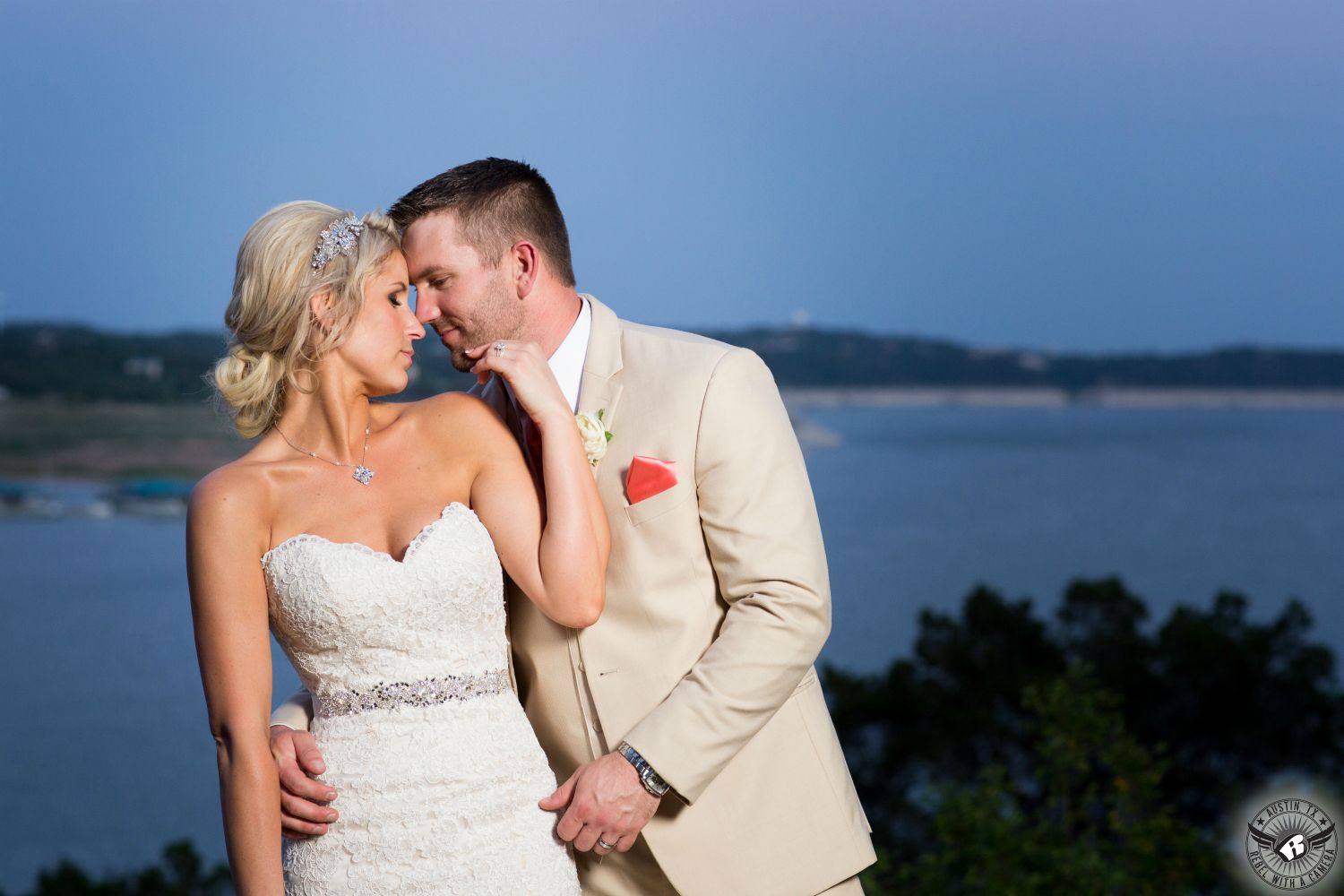 Bride in sparkly strapless wedding gown with diamond sash nuzzles groom in beige suit with amazing view of Lake Travis at Nature's Point wedding venue in Austin picture takes by Austin wedding photographer.