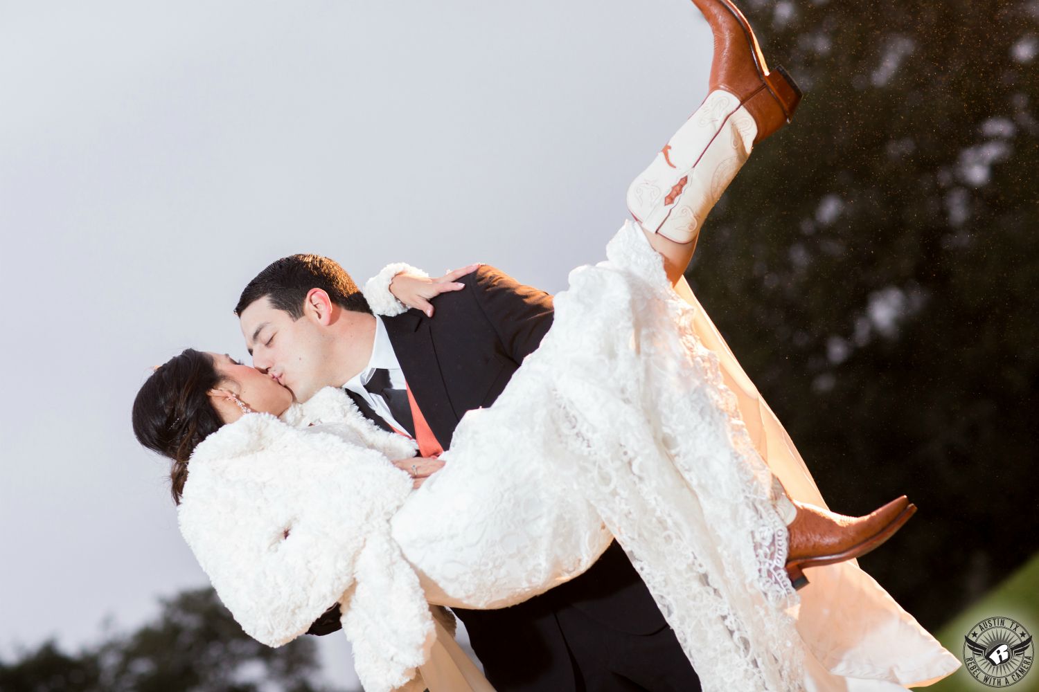 Groom in black tuxedo and orange vest holds bride in lace wedding dress and white fur coat as she kisses him and kicks her Longhorn boots in the air at Rustic wedding venue Gabriel Springs in Georgetown, Texas.