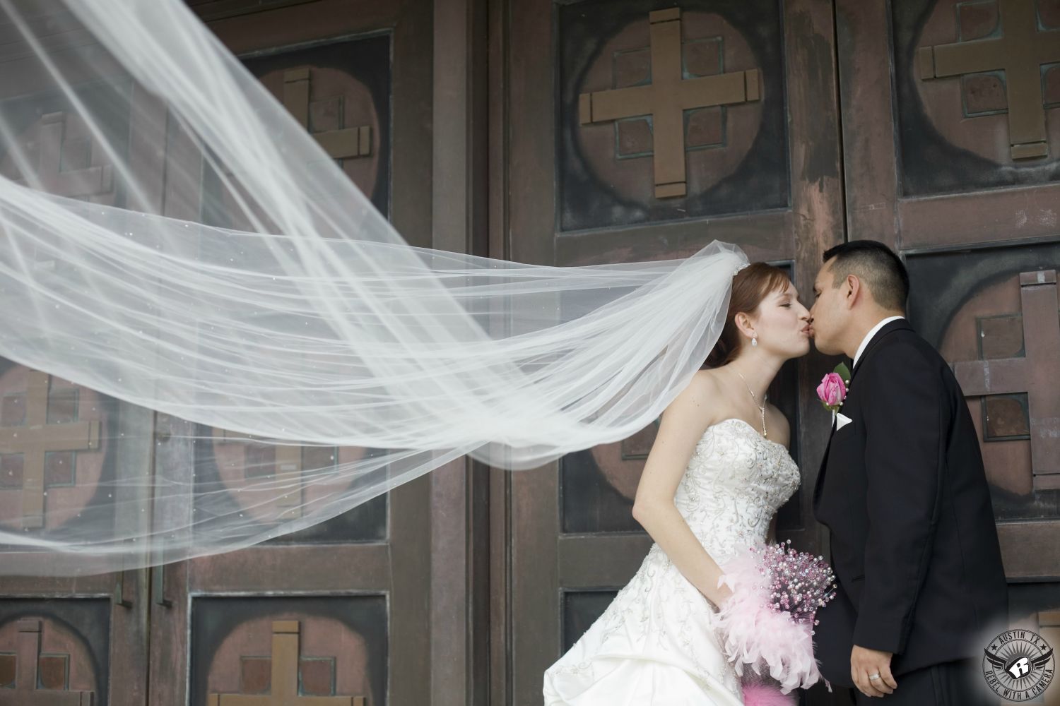 Brunette bride in sparkly white strapless wedding gown with pink feather and bling handmade bridal bouquet and groom with pink rose boutonniere kiss in front of bronze doors of Catholic church in Victoria, Texas, while her veil is lifted up across the frame.