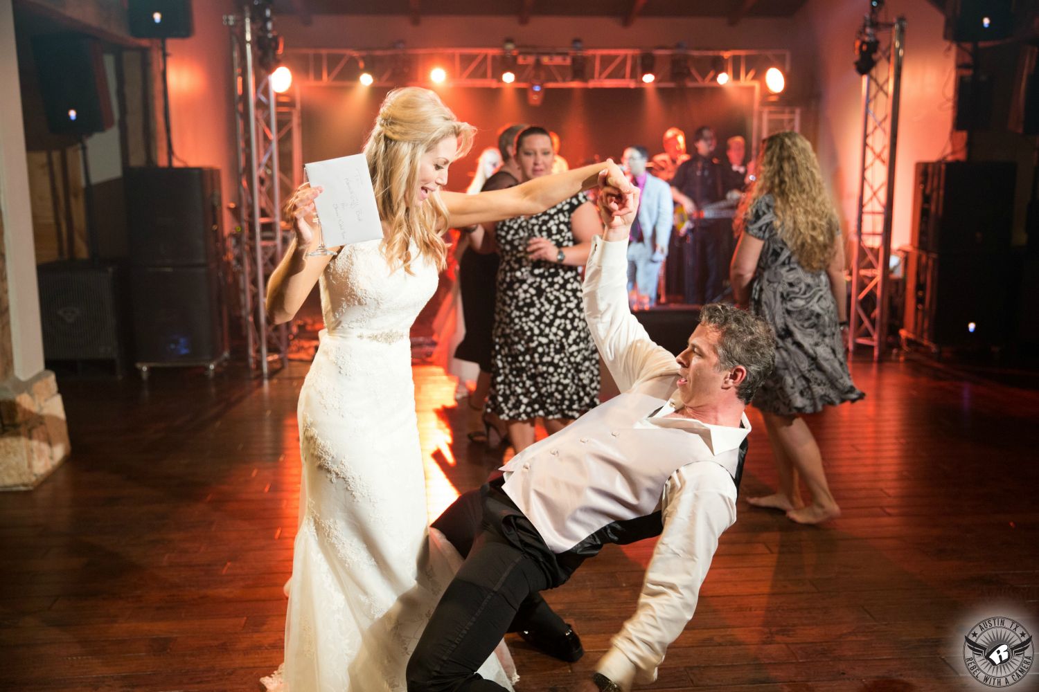 Wedding picture of groomin black tuxedo pants and white vest dancing with blonde bride with hair down at their wedding reception at Camp Lucy Dripping Springs wedding venue with fabulous orange stage lighting during wedding coordinated by Your Wedding, Your Way, Austin wedding Planner with the Pictures band playing on stage taken by wedding photographer in Austin.