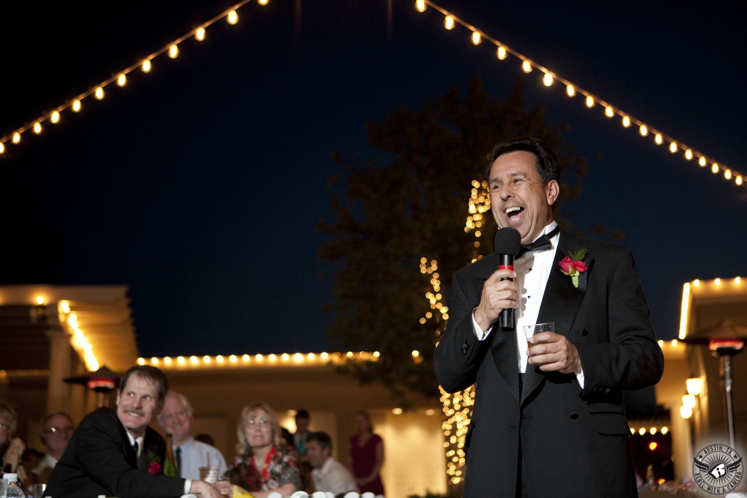 Happy father of the bride in black tuxedo and red rose boutonniere toasts couple at the wedding reception with twinkly lights strung behind him.