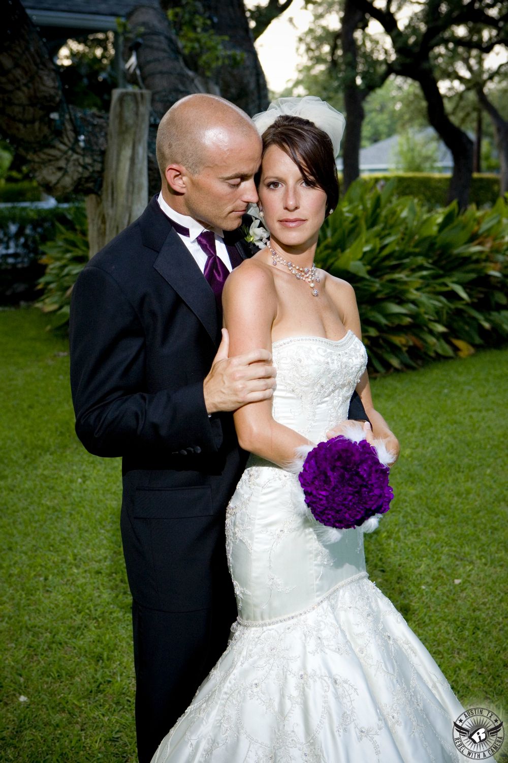 Groom with shaved head in black tuxedo and deep eggplant colored necktie nuzzles bride in elegant strapless mermaid wedding gown with purple carnation and white feather bridal bouquet at Green Pastures Austin wedding venue.