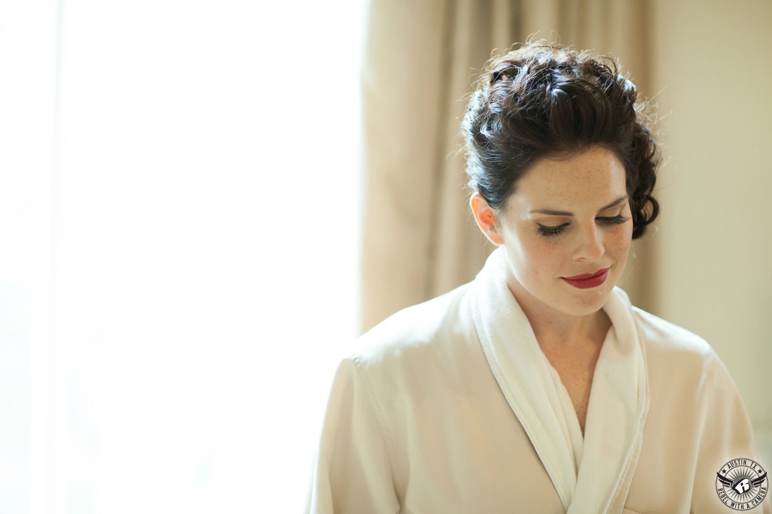 Dramatic picture of brunette curly haired bride with updo with gorgeous red lipstick on her lips hair and makeup by Mandy Hernandez looking down as she stands under the window in the bride's room at the Driskill Hotel in downtown Austin on her wedding day.