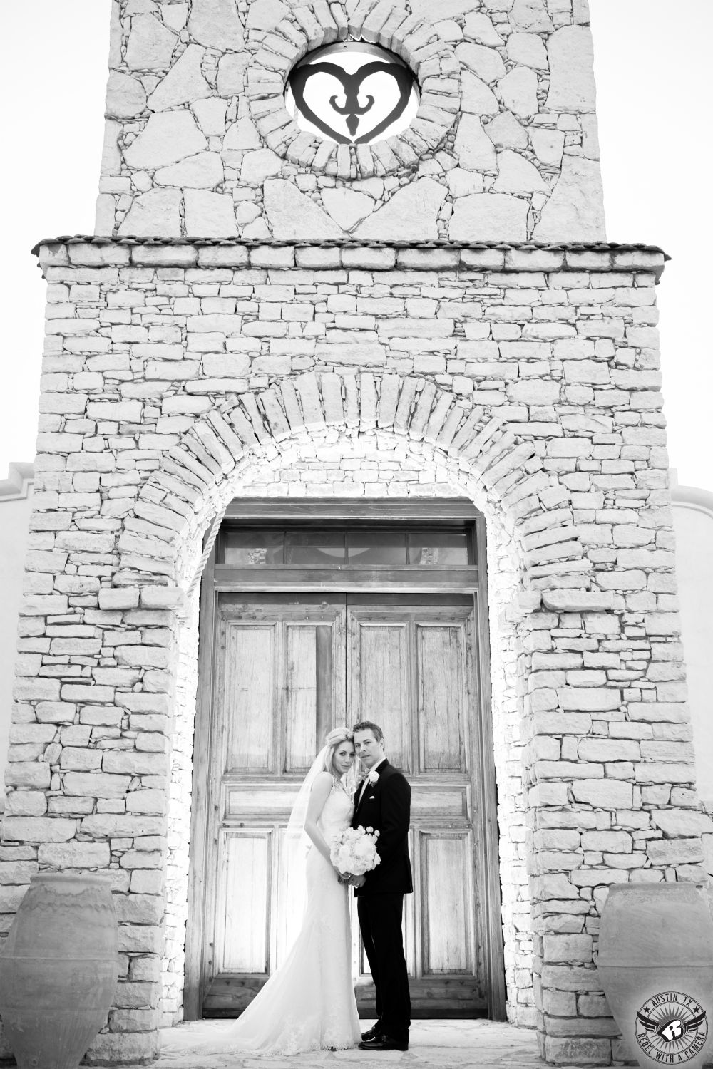 Dramatic black and white image of bride with delicate bouquet from Exquisite Petals Austin wedding florist and groom in black tuxedo stand in front of the wooden doors of Ian's Chapel at Camp Lucy the wedding venue in Dripping Springs, the wedding capital of Texas wedding coordinated by Your Wedding, Your Way, Austin wedding Planner.