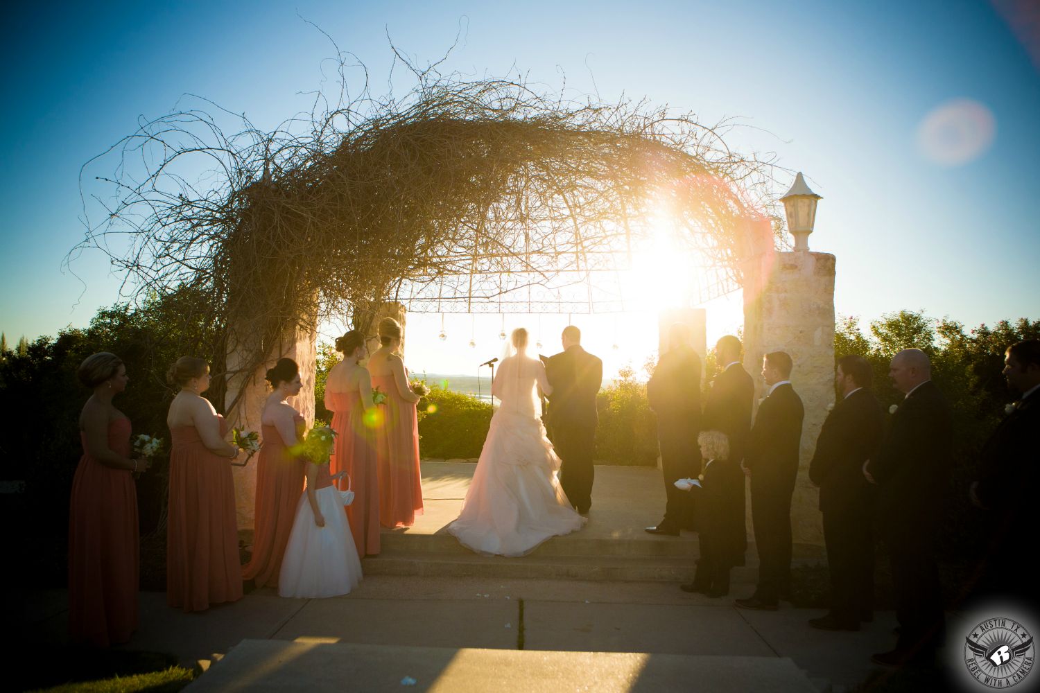 Austin wedding photographers take wedding picture of gorgeous wedding ceremony with bridesmaids in pink dresses and bride in pale pink wedding dress at the outdoor arbor wedding ceremony site at Vintage Villas looking out over Lake Travis with the sunset behind.
