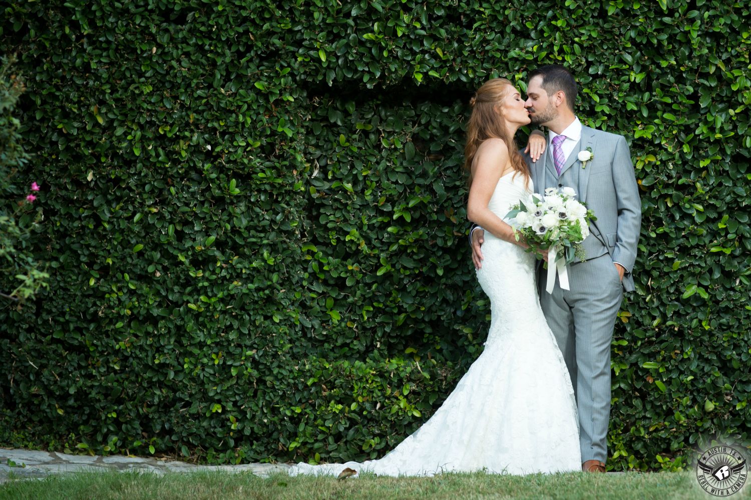 Austin wedding photography of groom in grey suit who is kissing gorgeous, tall, red head bride who had wedding makeup done by Makeup by Adrienne in front of fig ivy covered wall at Nature's Point Austin wedding venue.