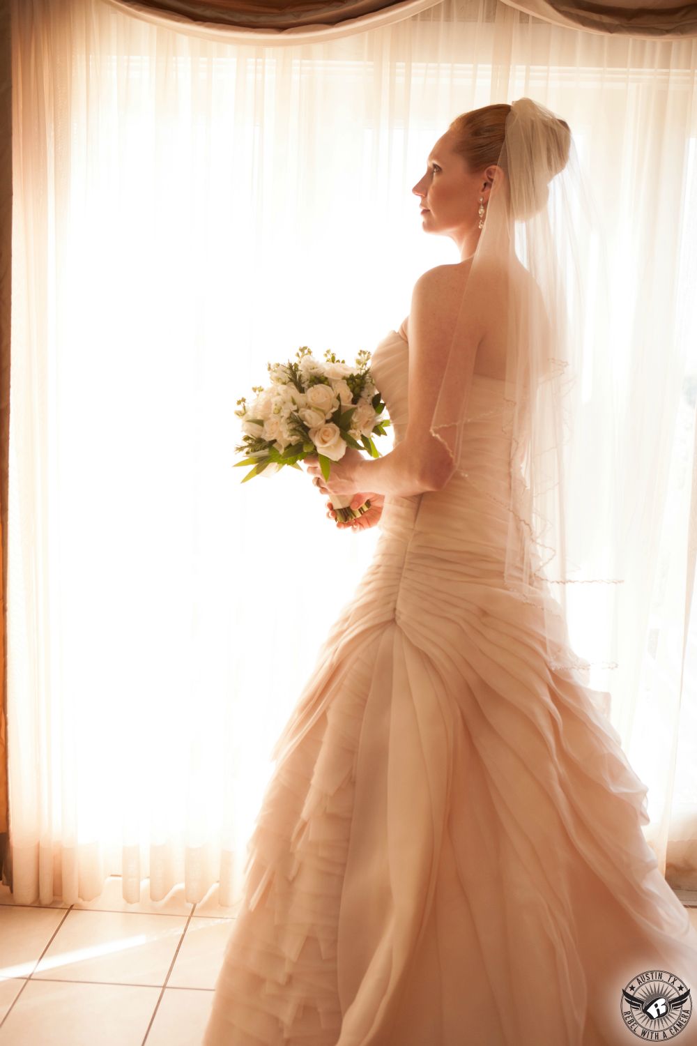 Austin wedding photography of bride in elegant, light pink, ruffled wedding dress with long bridal veil with bridal bouquet by Verbena Floral Design Austin wedding florist stands in front of window in the bride's room at Vintage Villas wedding venue near Lake Travis.