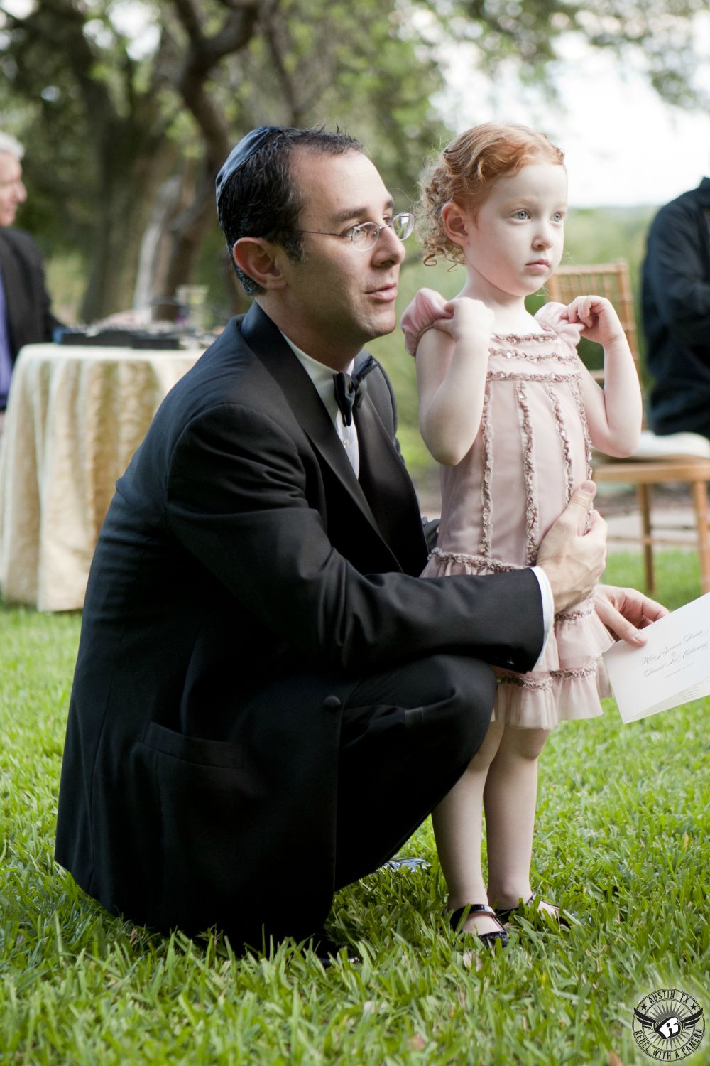 Dad in black tuxedo and black bowtie watches Jewish outdoor wedding ceremony at the Four Seasons Hotel in Austin by Lady Bird Lake with adorable little girl in delicate pink dress. 