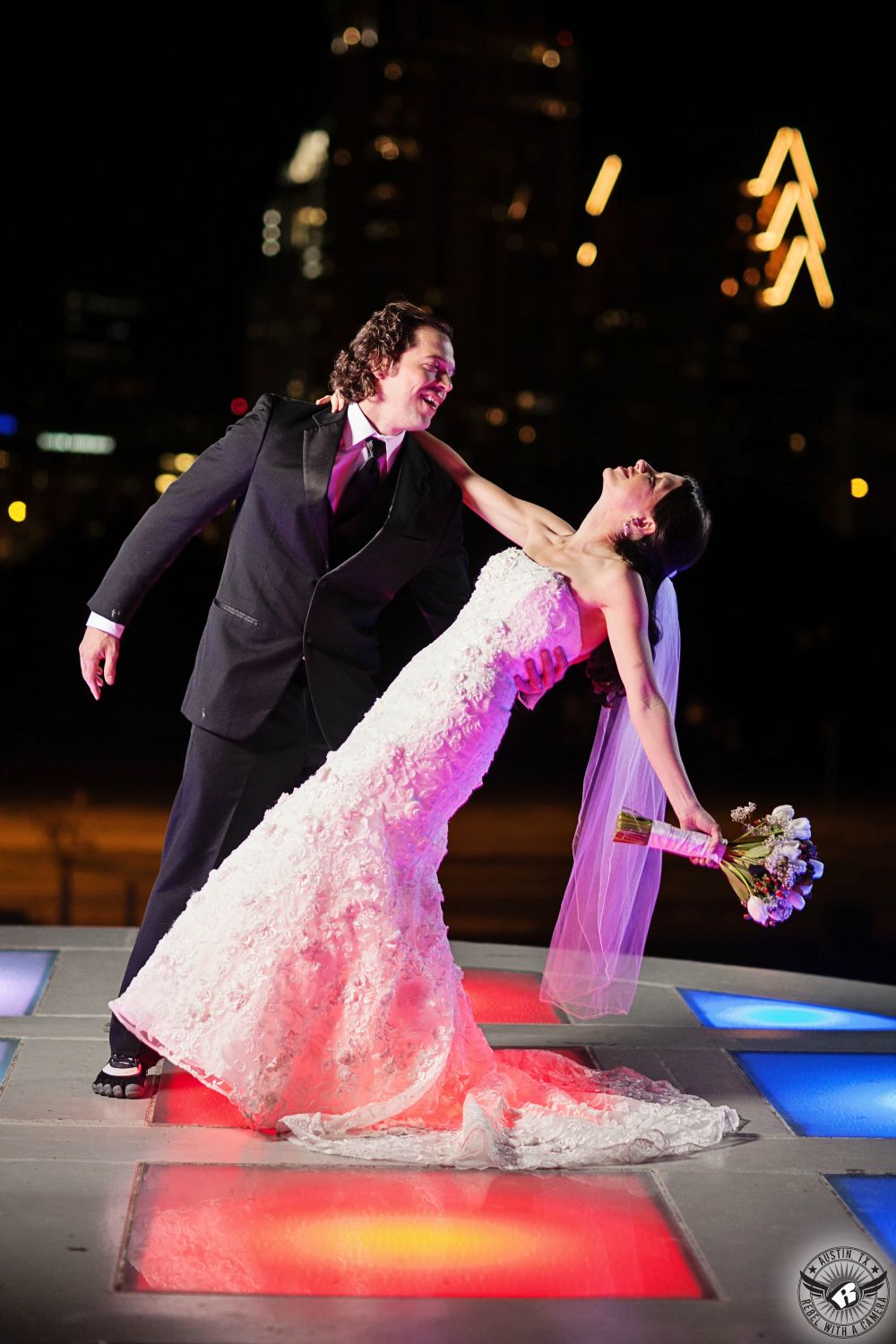 Austin wedding photography of groom in black tuxedo dipping bride in beaded white strapless wedding dress on colored light platform at the Long Center with the Austin skyline in the back at night during their wedding day.