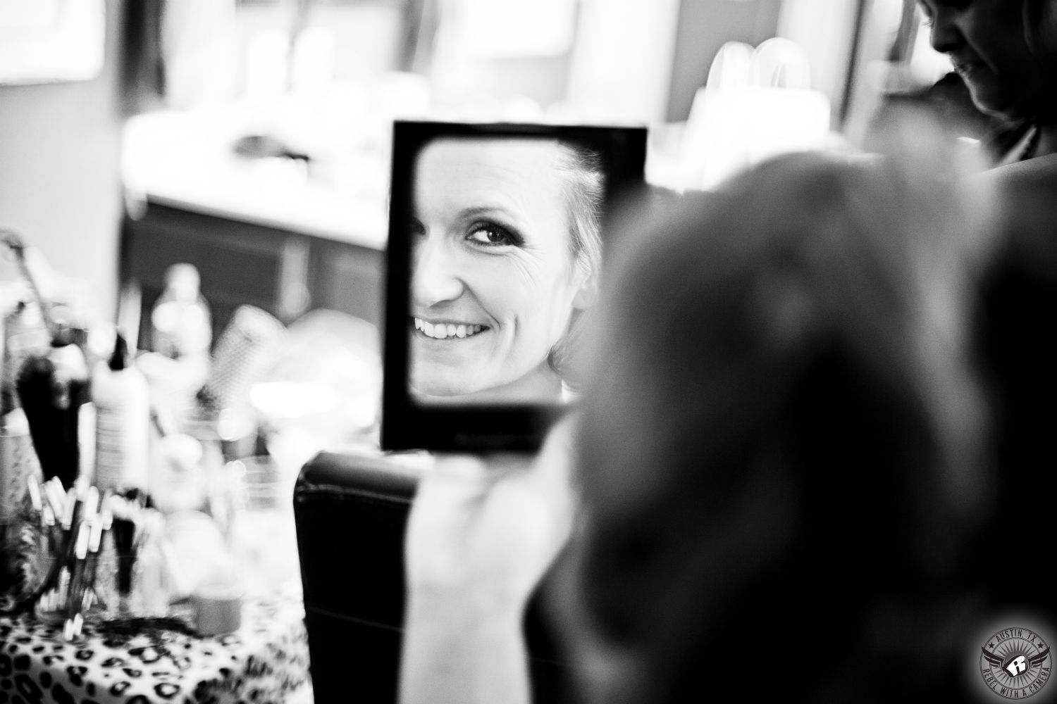 Black and white image of happy bride looks at her reflection in mirror in the bride's room on her wedding day while she gets married with hair and makeup by Divaz Fabula, Austin wedding makeup and hair stylists.