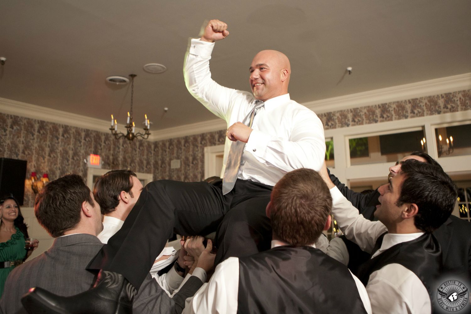 Groom with shaved head in white dress shirt and grey tie is lifted on the shoulders of the guests pumps fist in the air during the wedding reception at Green Pastures the wedding venue in South Austin.