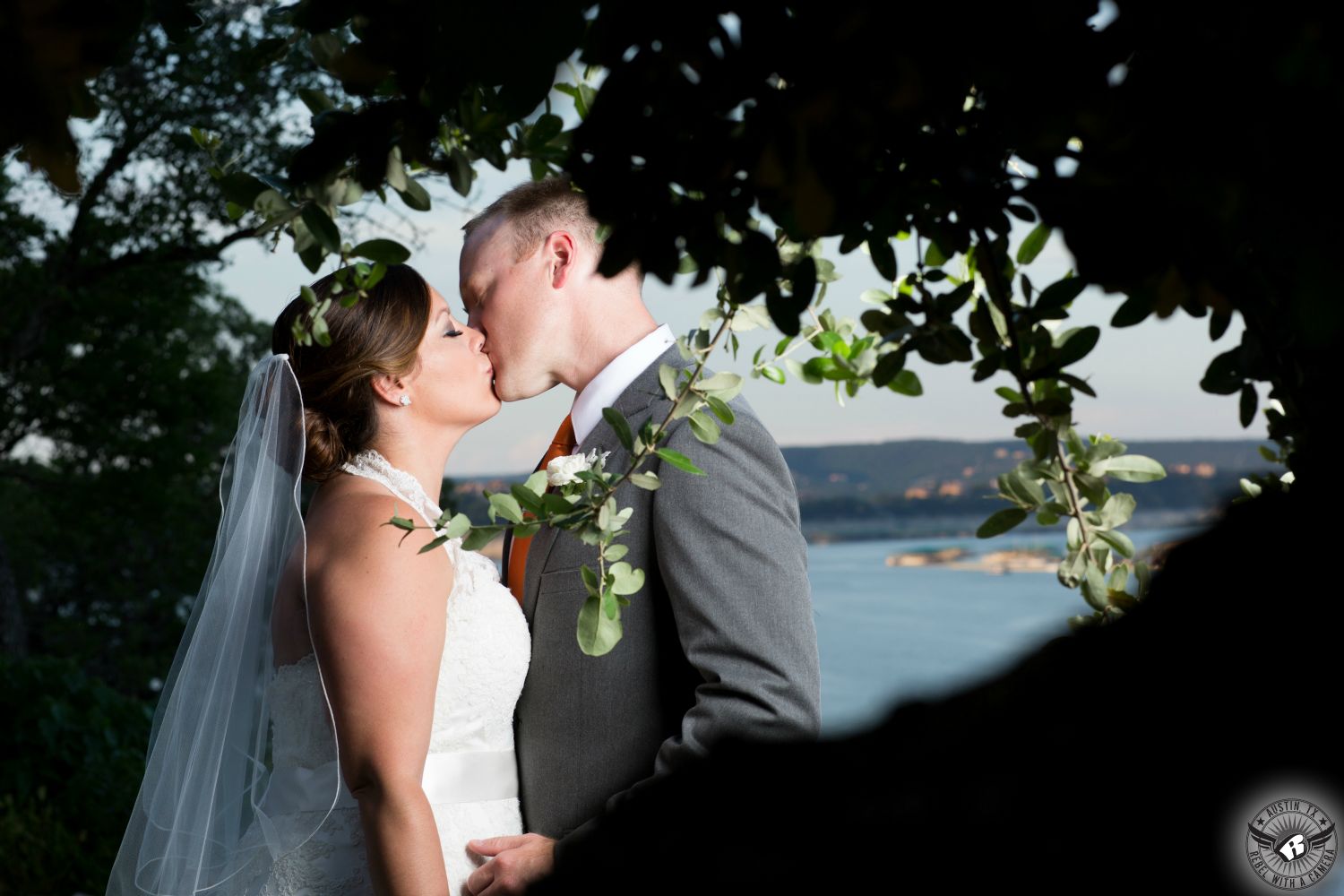 Fabulous Austin wedding photography of bride with hair and makeup by Urban Betty and groom in grey suit with boutonniere by Bouquets of Austin wedding florist kissing inbetween leaves on a tree overlooking Lake Travis at the Central Texas wedding venue, Nature's Point.