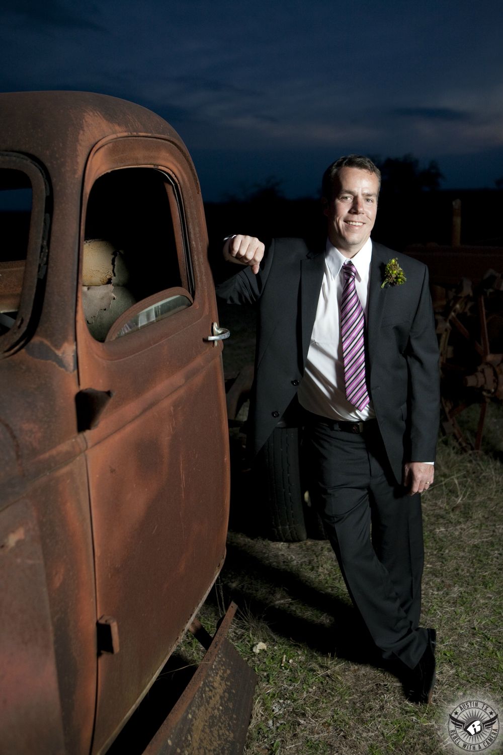 Dapper groom in pink and purple striped tie and black suit leans against rusted old truck on his wedding day at the Hill Country wedding venue, Star Hill Ranch.