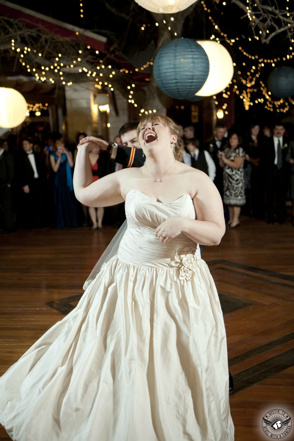 Super happy bride in gorgous strapless ballgown wedding dress is laughing joyously as groom in military dress blues uniform twirls her on the dance floor under paper lanterns hanging from the trees with twinkly lights at Kindred Oaks the Austin wedding venue while Dexter Kyner DJ for Hire spins some tunes.