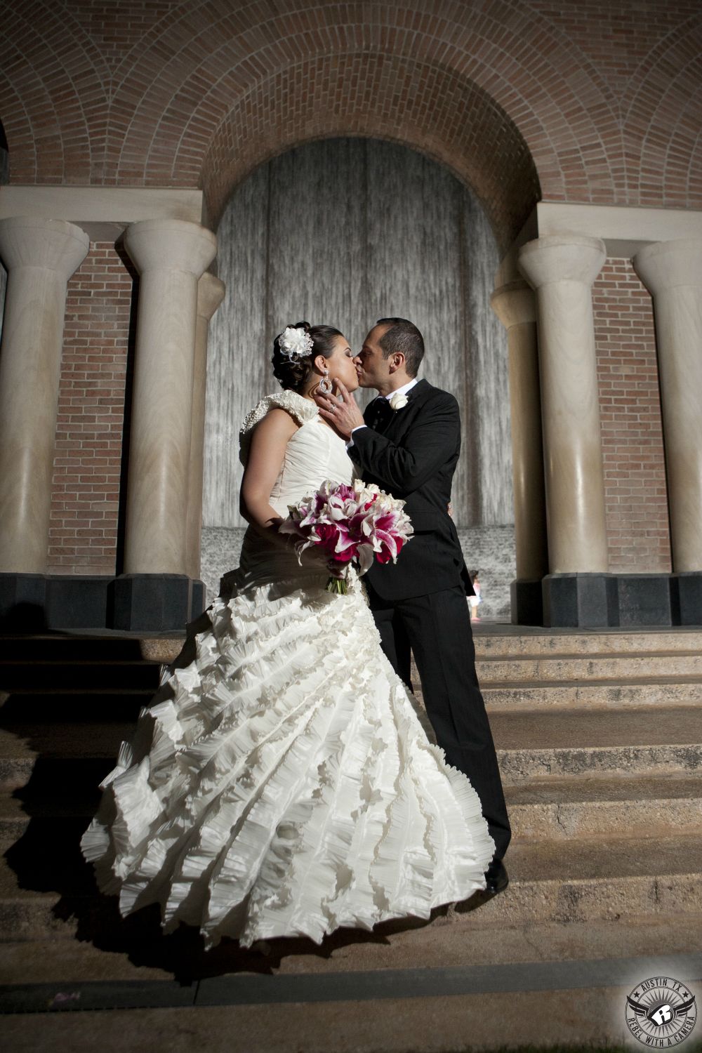 Bride in one shouldered ruffled wedding dress with stunning magenta stargazer lily bouquet by Weddings in Bloom Houston wedding florist and groom kiss passionately in front of the water wall in Houston during their Lebenese wedding at the Westin Galleria.