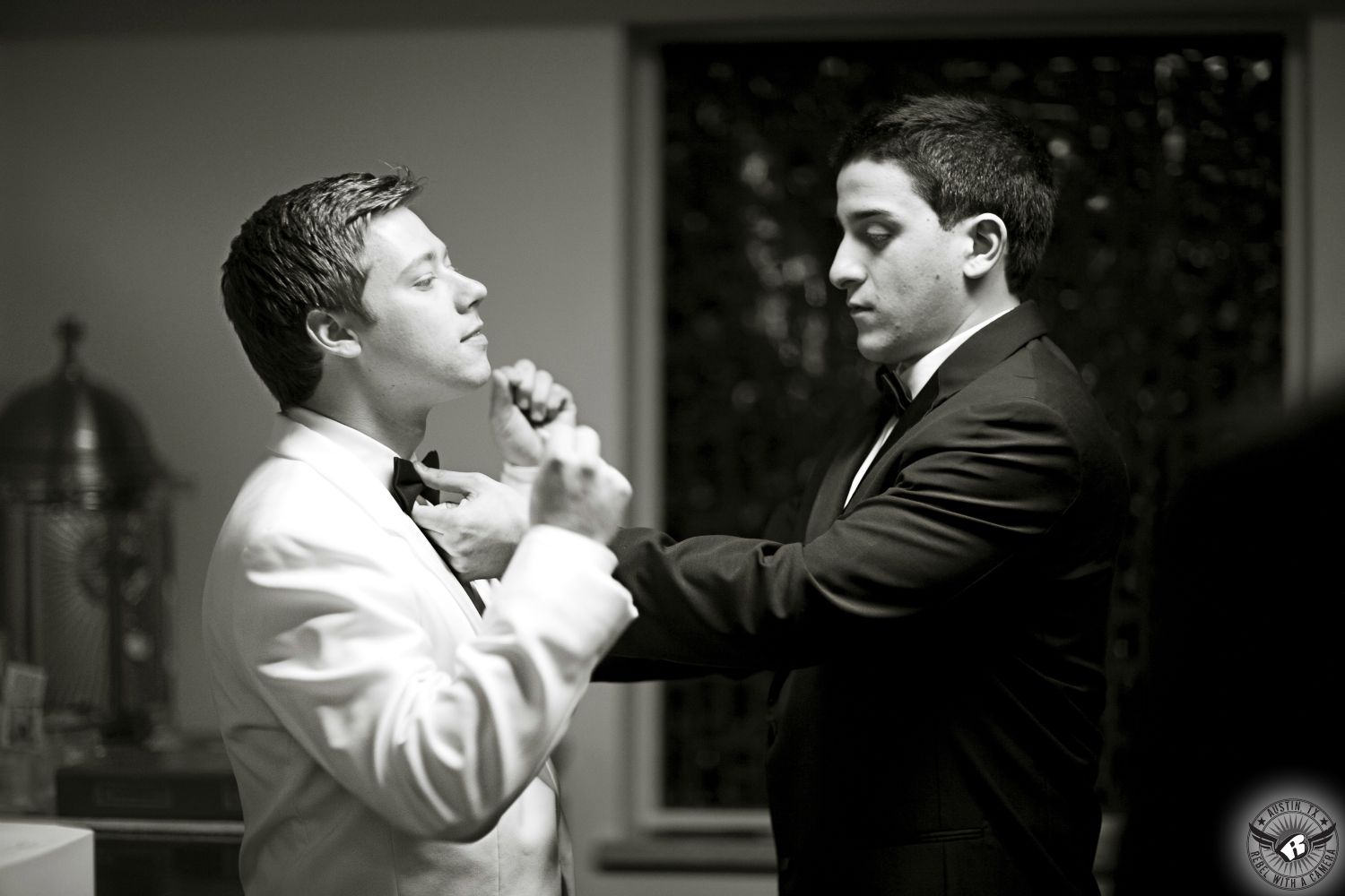 Best man in black tuxedo helps adjust the black bowtie of the groom in a classic white tuxedo jacket the at St. Austin's before the Catholic wedding ceremony.  