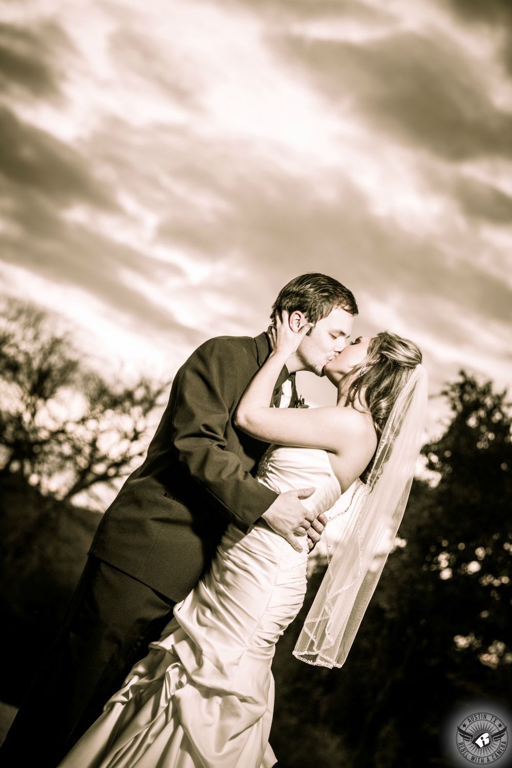 Sepia toned image of bride and groom kissing passionately with an amazing sunset behind them at Gabriel Springs Central Texas wedding venue taken by Austin wedding photographer by Rebel with a Camera.