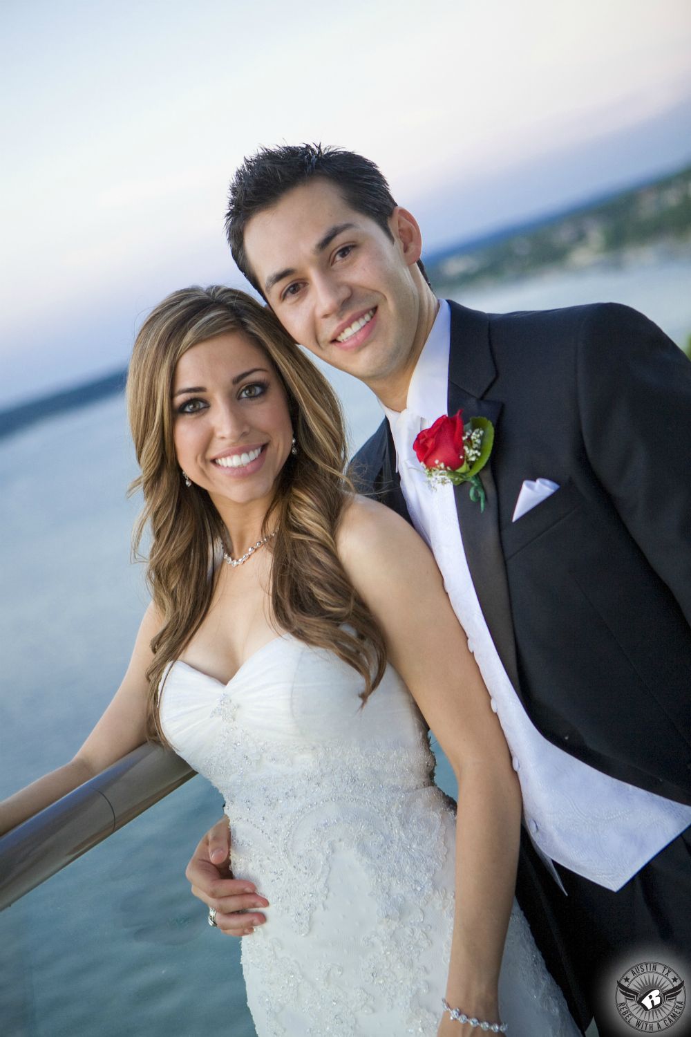 Very happy bride in strapless bridal gown and groom in black tuxedo with red rose boutonniere pose on the deck on their wedding day outside the Lakeway Resort and Spa Austin wedding venue.