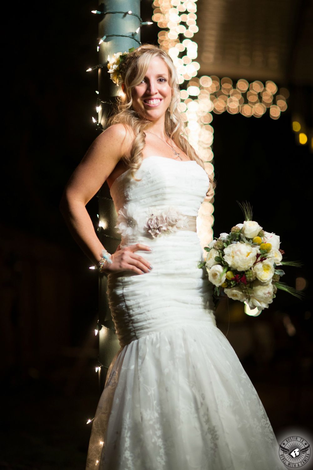 Austin wedding photography of stunning, tall, blonde bride in strapless mermaid bridal gown posing for picture and holding stunning bouquet by Magpie Blossoms with lots of twinkly lights at Sicola's Garden House.