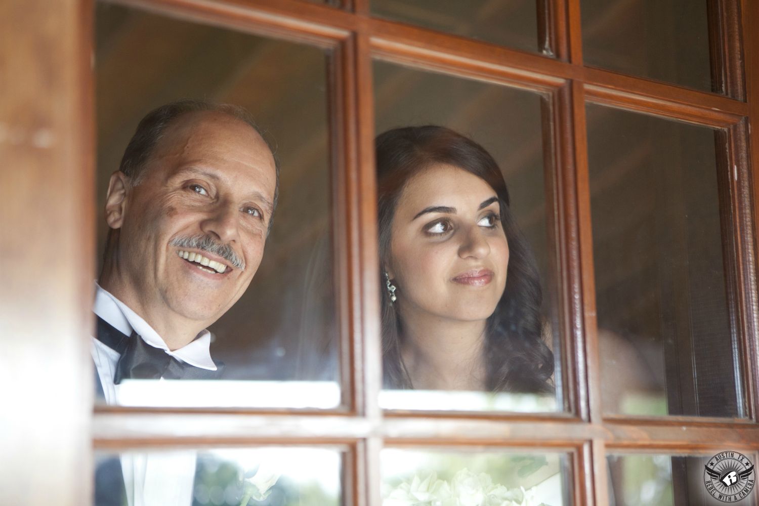 Bride and her father in black tuxedo look out of the window just before he walks her down the aisle to the wedding ceremony at the Grande Hall at Hofmann Ranch wedding venue in San Antonio, Texas.
