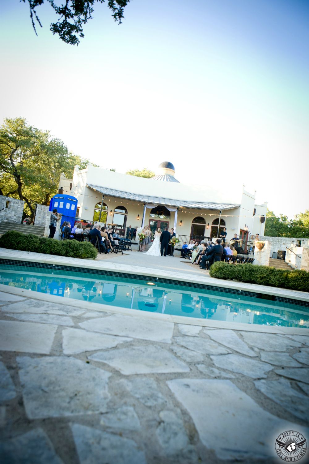 Wide shot of outdoor wedding ceremony site from across the pool at Hamilton Twelve Mediterranean wedding venue near Austin, Texas with Dr. Who TARDIS in the background.
