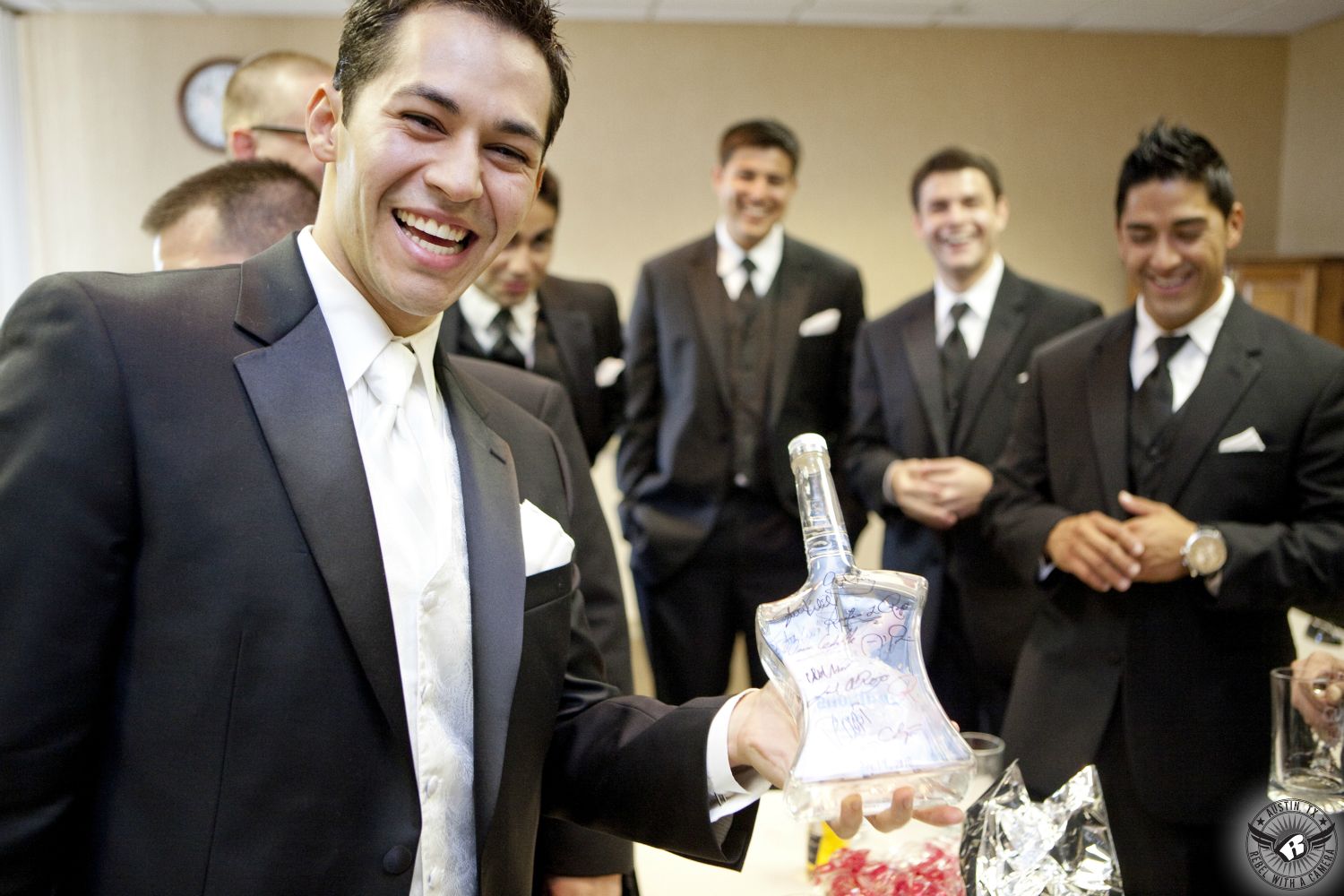 Picture of happy groom in black tuxedo and white vest and tie showing off bottle of specialty alcohol he received as a gift on his wedding day with his groomsmen in the groom's room at the Queen of Angels Chapel in Spicewood, Texas taken by Austin wedding photographer.
