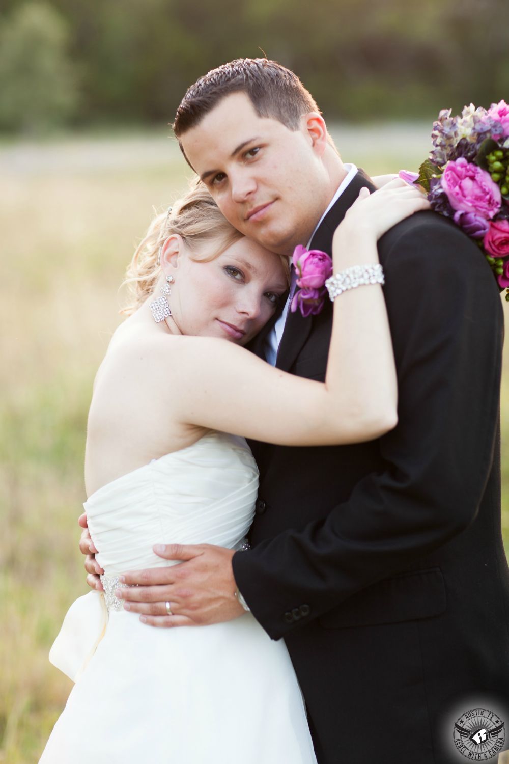 Blonde bride with intense gaze and magenta bouquet embraces groom with pink boutonniere in a field at Texas Old Town before getting married at Tejas Hall.