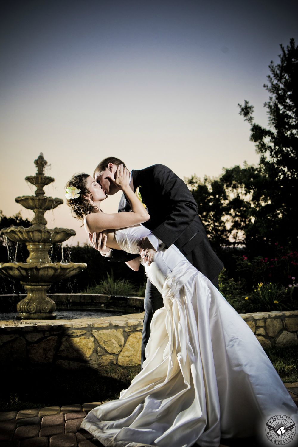 Groom dips bride in elegant strapless mermaid wedding dress with orchid in her hair and kisses her in front of fountain at Nature's Point the Austin wedding venue on the shores of Lake Travis.
