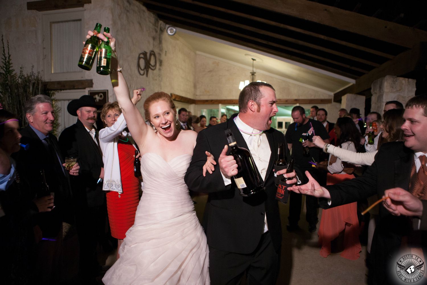 Wild tall red headed bride in pale pink ruffled wedding dress and groom in black tuxedo carry multiple bottles of alcohol and excitedly exit their wedding reception at Vintage Villas on Lake Travis. 