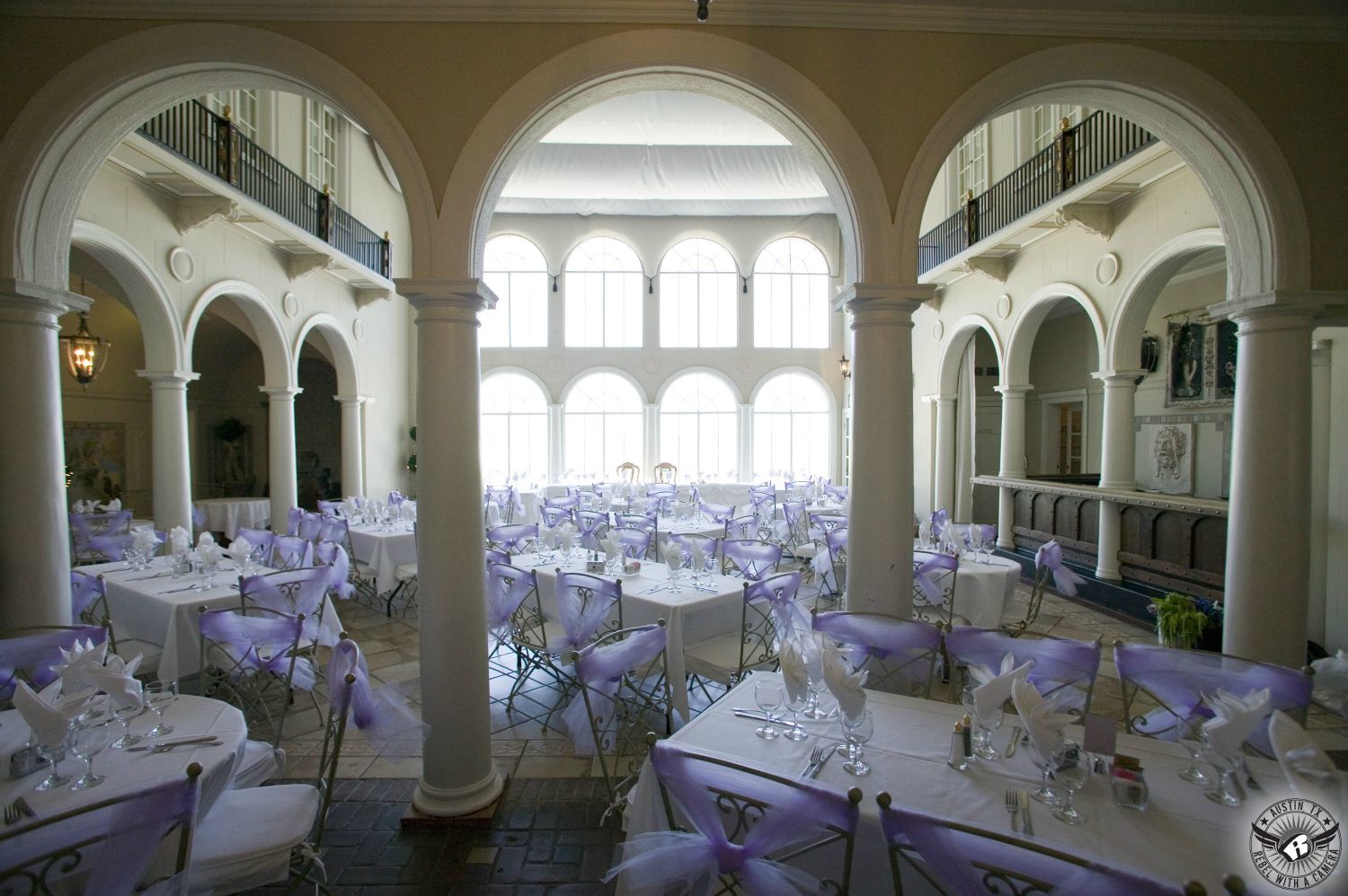 Picture of unbelievably stunning wedding reception venue with lots of windows, arches, and elegantly set tables with lavender chair ties taken by Austin wedding photographer.