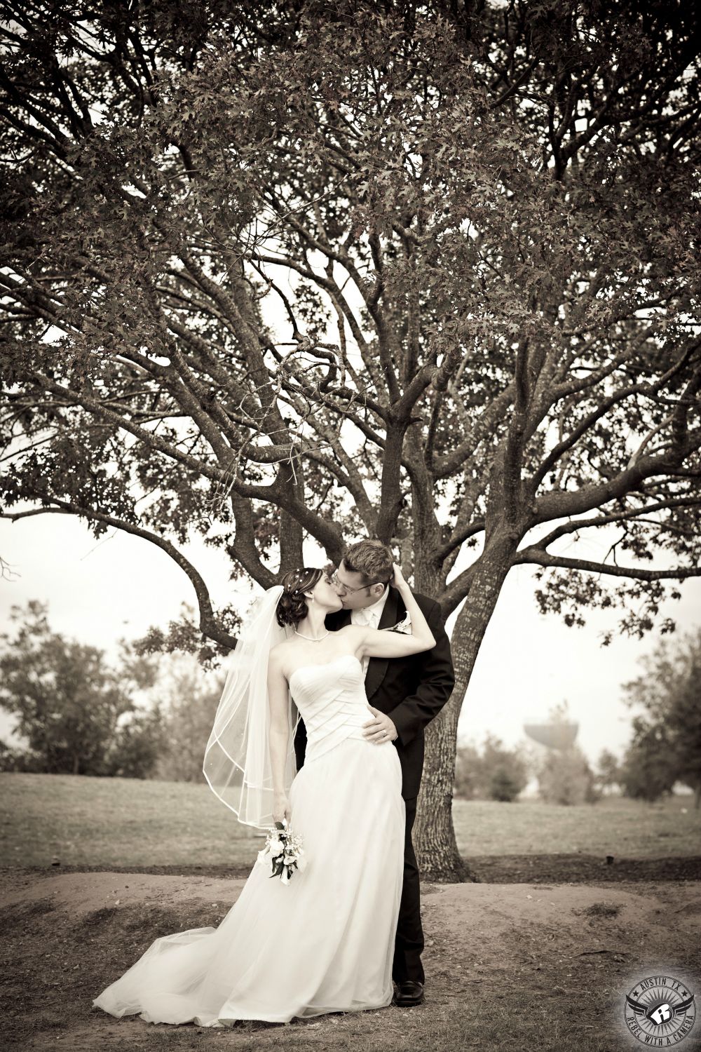 Bride and groom kiss passionately in front of beautiful tree on their wedding day at Mueller Lake Park in Austin, Texas.