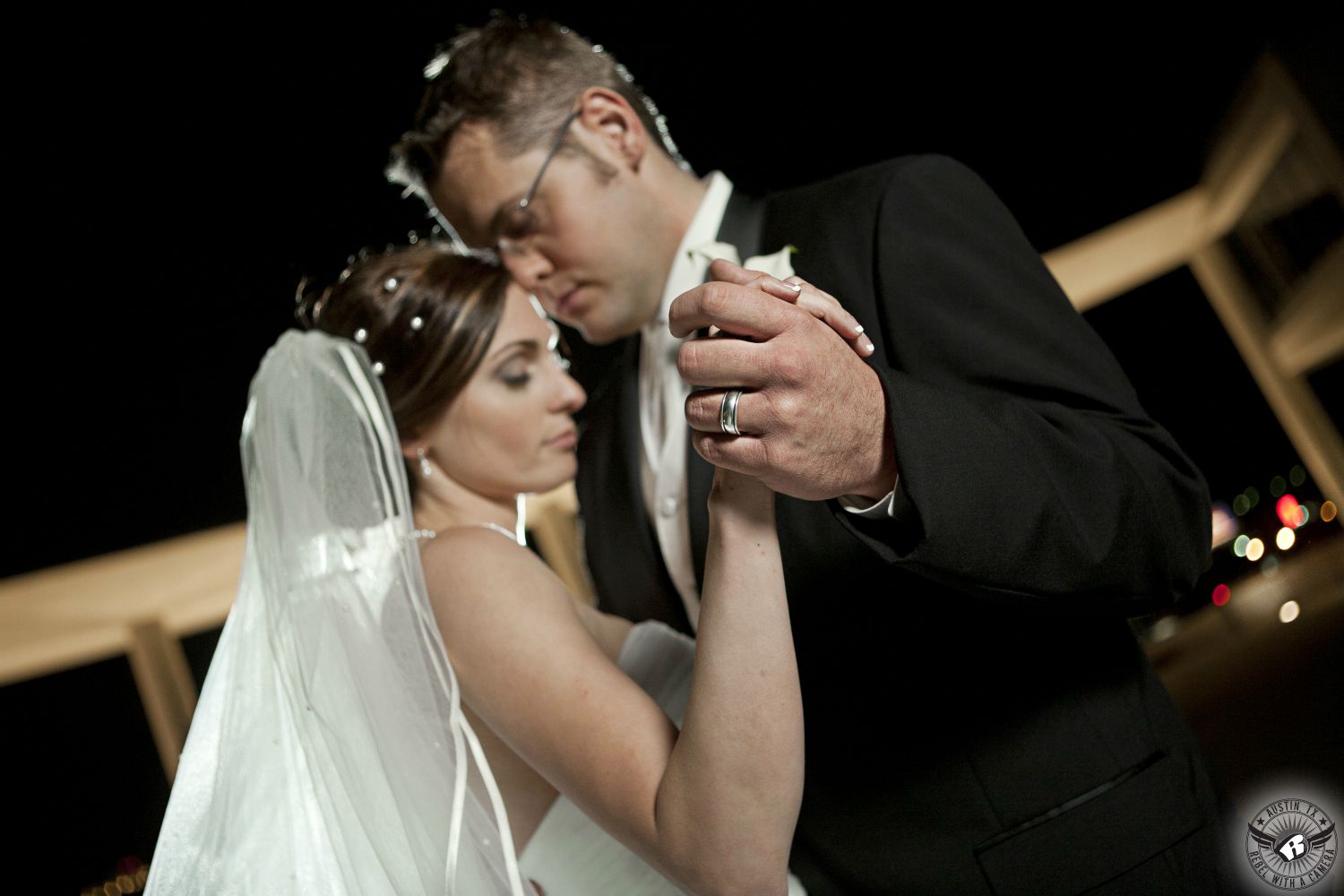 Bride in strapless dress, veil, and pearls in her hair and groom in black tuxedo and white vest and tie dance at night at the Long Center in Austin, Texas, on their wedding day with the groom's wedding ring in focus and the couple being backlit.