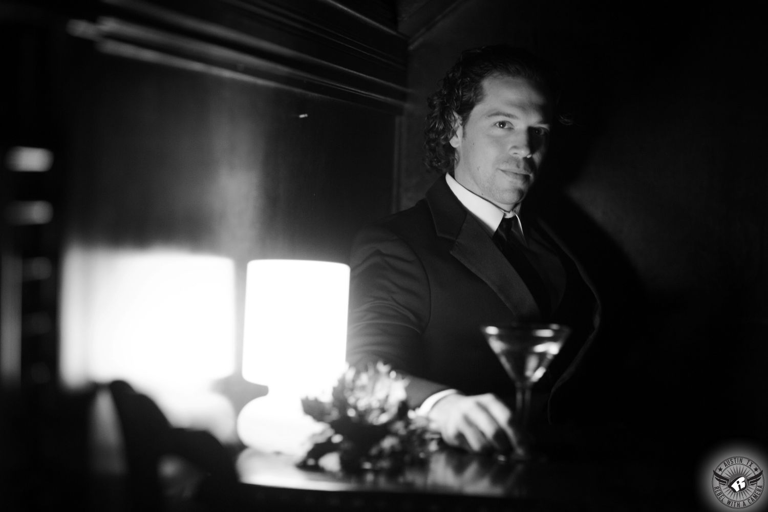 Black and white image of groom in black tuxedo and curly hair and martini glass looking straight at the camera as he poses in a relaxed manner with dramatic light in the bar at Green Pastures Austin wedding venue.