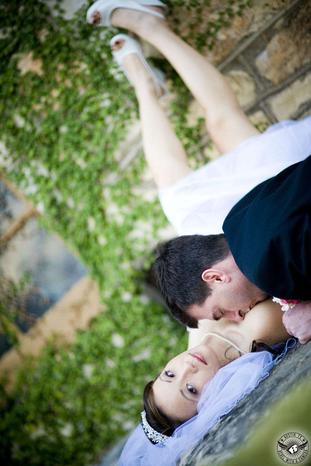At Nature's Point wedding venue near Austin, Rebel with a Camera, Austin wedding photographer, takes picture of bride reclining on her back with legs up on fig ivy covered wall while groom kisses her neck. 