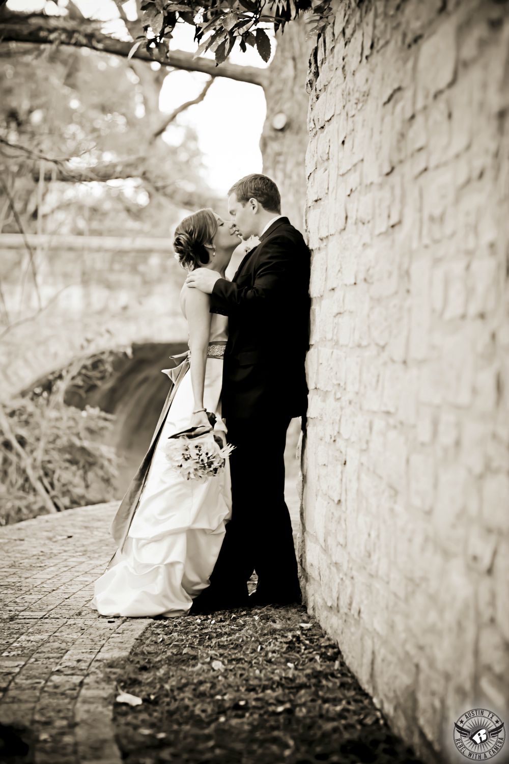 Sepia toned image of groom kissing bride in ruffled strapless wedding gown as he leans against high stone wall at the Etter-Harbin Alumni Center wedding venue on the University of Texas campus.