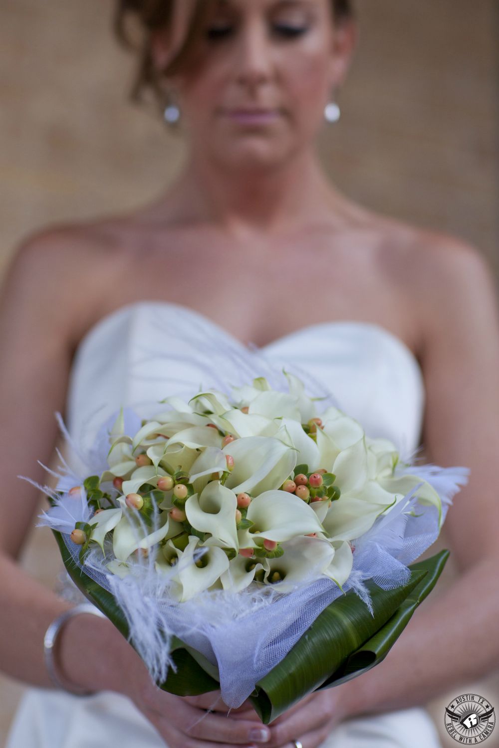Gorgeous bridal bouquet by the Glass Fountain Austin wedding florist with white calla lillies, white feathers, and yellow orange Hypericum Berries held by the bride in a strapless wedding dress at the Etter-Harbin Alumni Center wedding venue on the University of Texas campus.