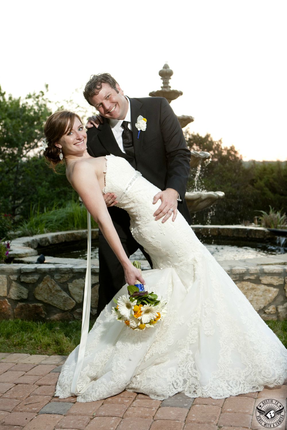 Bride in strapless lace mermaid styled dress with daisy and yellow billy balls bridal bouquet by Bouquets of Austin and makeup by Maris Malone Calderon and groom smile at the camera in front of camera at Nature's Point the Austin wedding venue.