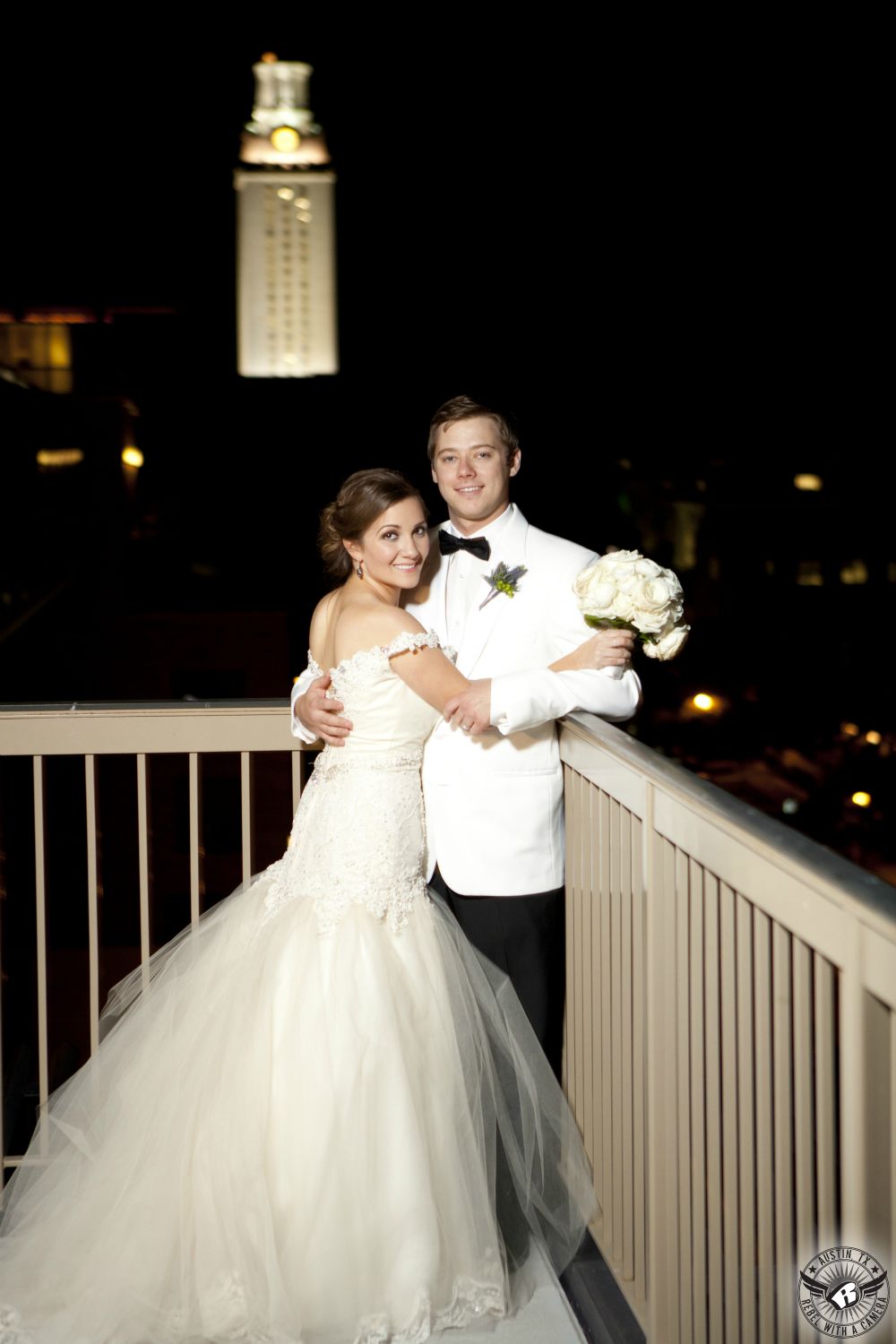 Bride in gorgeous wedding dress and groom in white tuxedo jacket and black bowtie happy in love standing on the balcony at the AT&T Executive Education and Conference Center at night with the UT tower in the background on their wedding day.