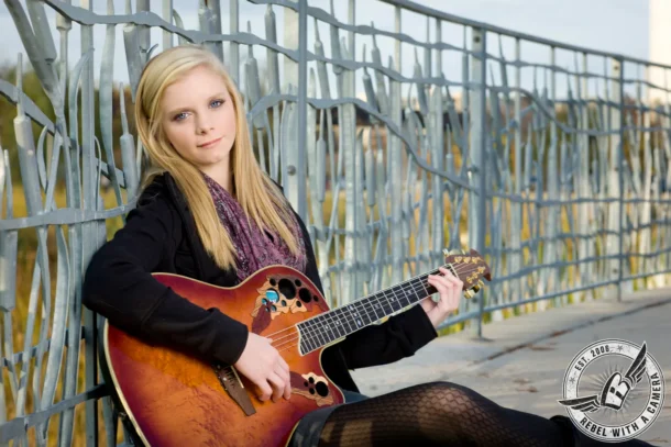 Austin senior portrait of a blonde woman holding an acoustic guitar beside a metal fence in Austin, TX.