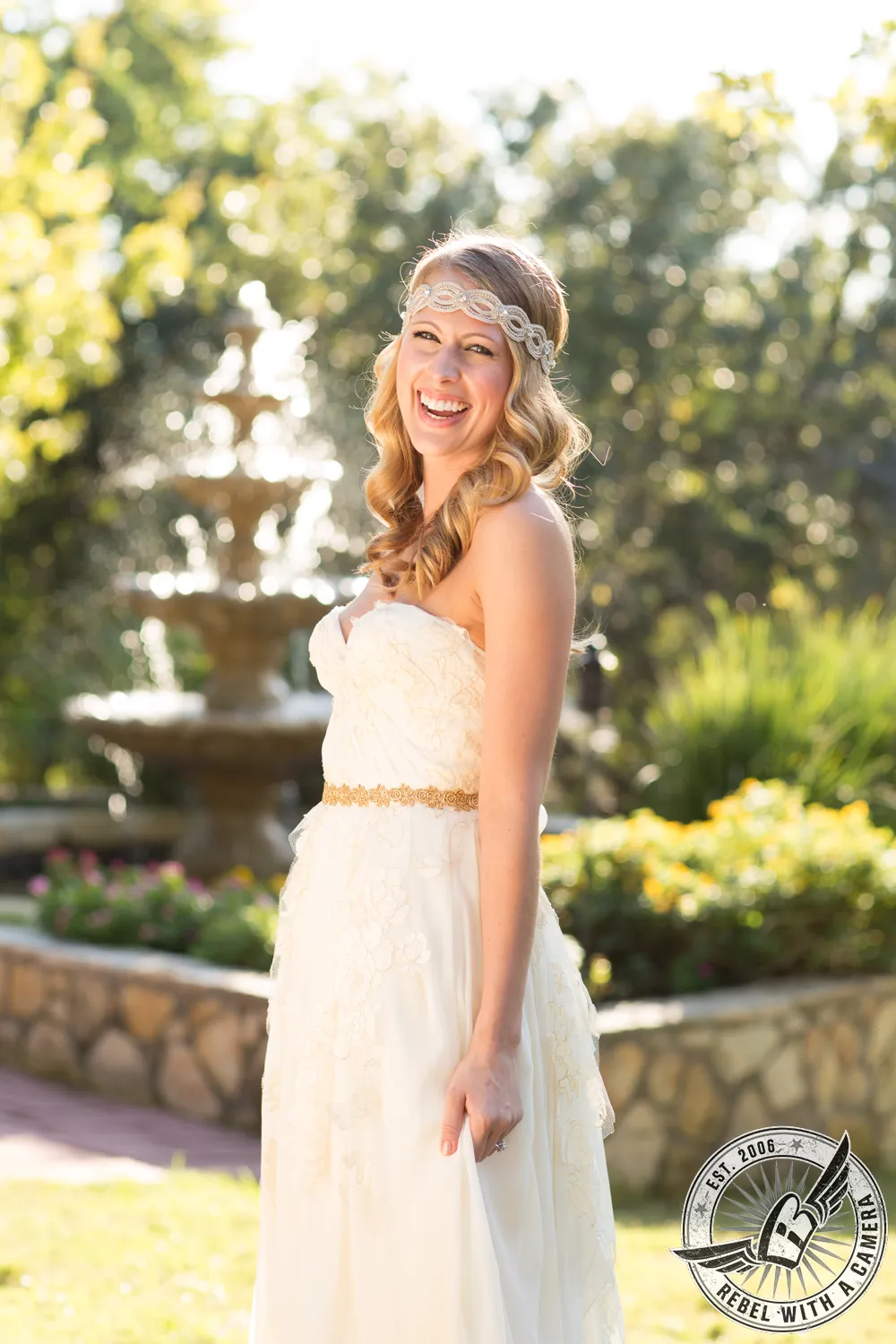 Smiling bride in a white wedding dress and jeweled headband standing beside a garden fountain in Austin, TX.