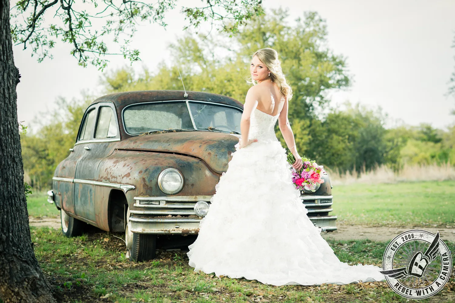 Bride in a white wedding dress holding a colorful bouquet beside a vintage car