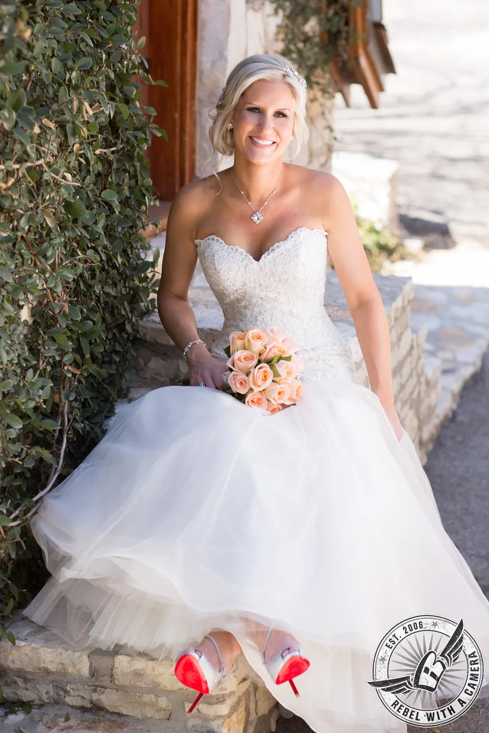Smiling bride in a white wedding dress holding a peach rose bouquet beside a hedge in Austin, TX.