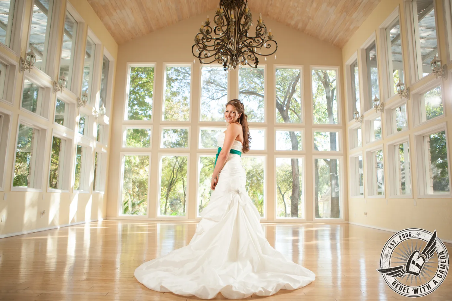 Bride in a white gown with a green sash standing in a bright glass-walled room beneath a chandelier in Austin, TX.