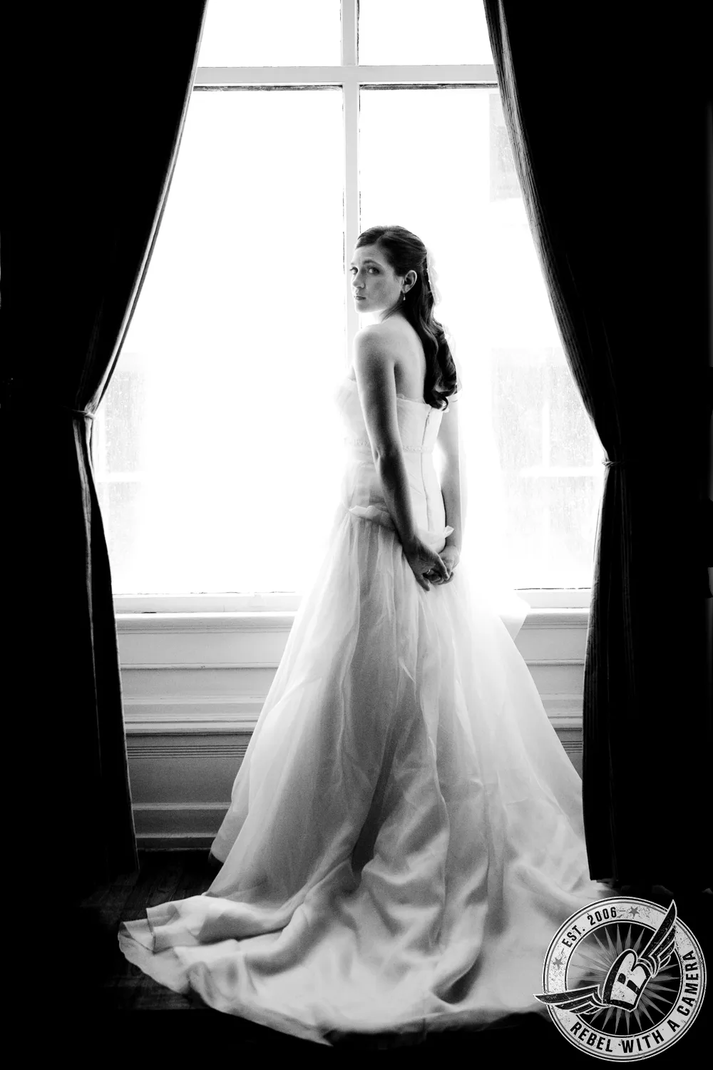 Black-and-white portrait of a bride in a flowing gown standing before a bright window framed by curtains in Austin, TX.