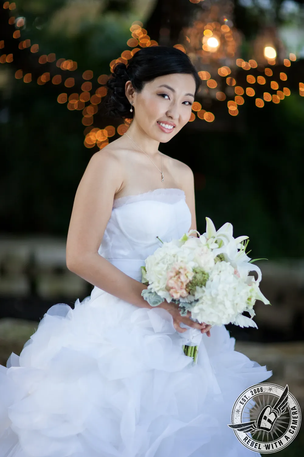 Bride in a strapless white dress holding a large bouquet with warm lights glowing behind her in Austin, TX.