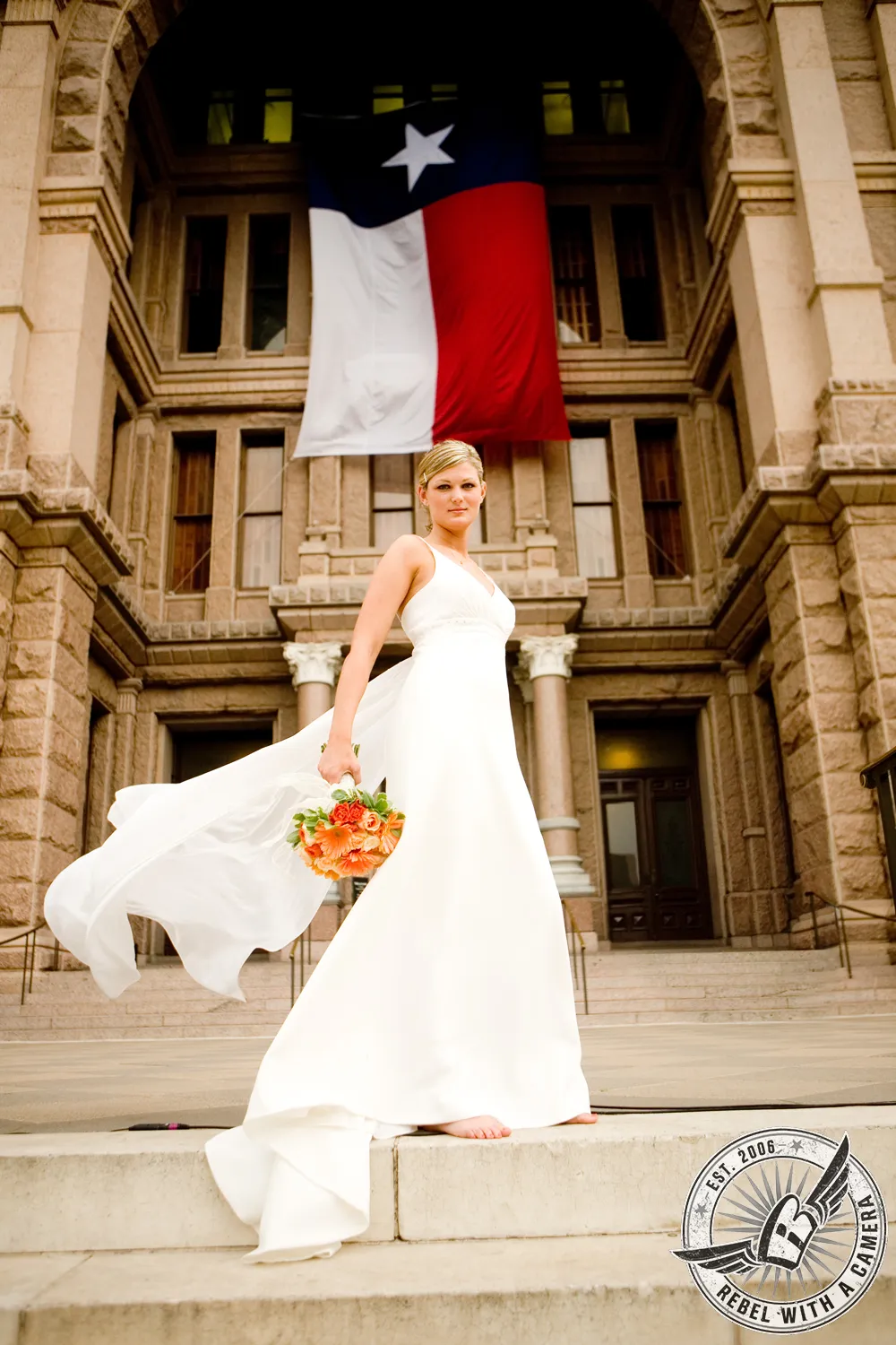 Bride in a white wedding dress holding an orange bouquet in front of the Texas flag and a historic building in Austin, TX.