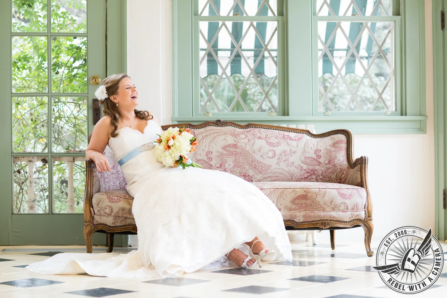 Laughing bride seated on a vintage floral sofa holding a bouquet in a bright room