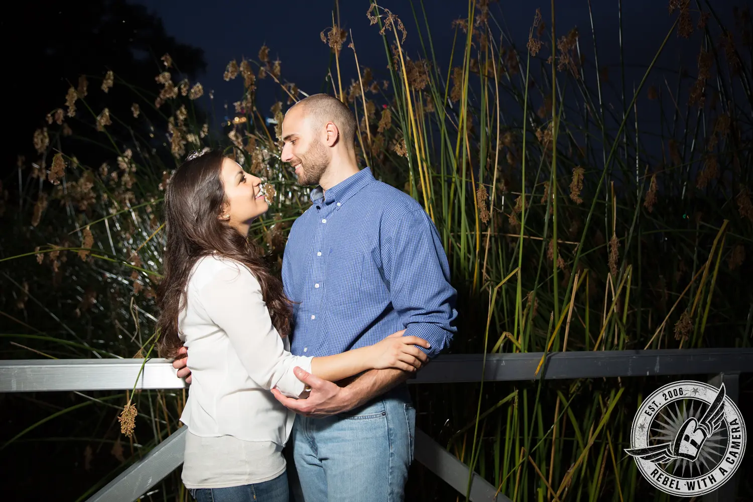Couple embracing on a bridge beside tall reeds at night in Austin, TX.