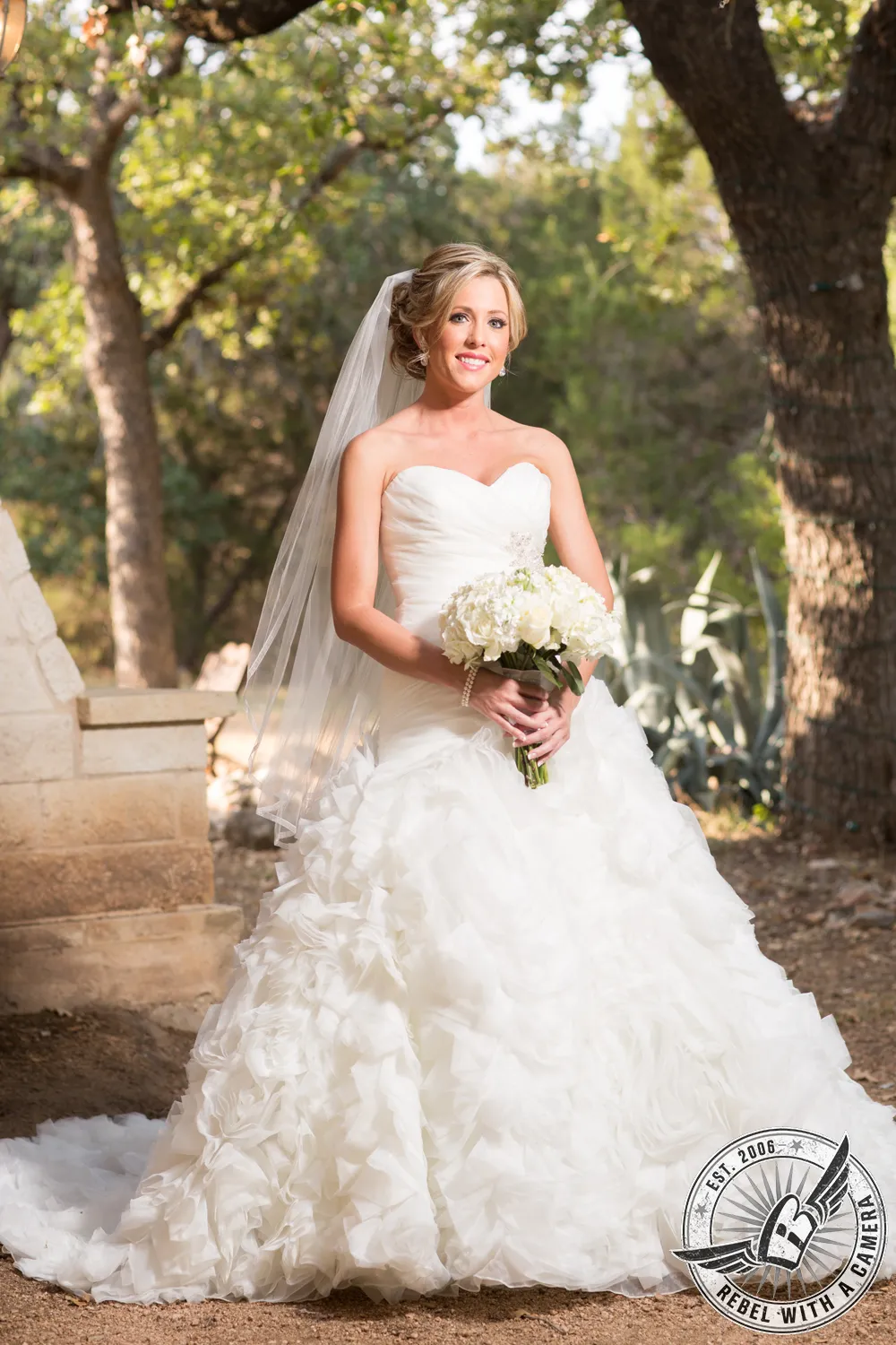 Bride in a ruffled wedding dress holding a white bouquet in a wooded setting in Austin, TX.